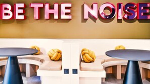 Ein Zimmer mit einem Wandschild mit der Aufschrift "BE THE NOISE" und zwei runden Tischen mit gelben Kissen.