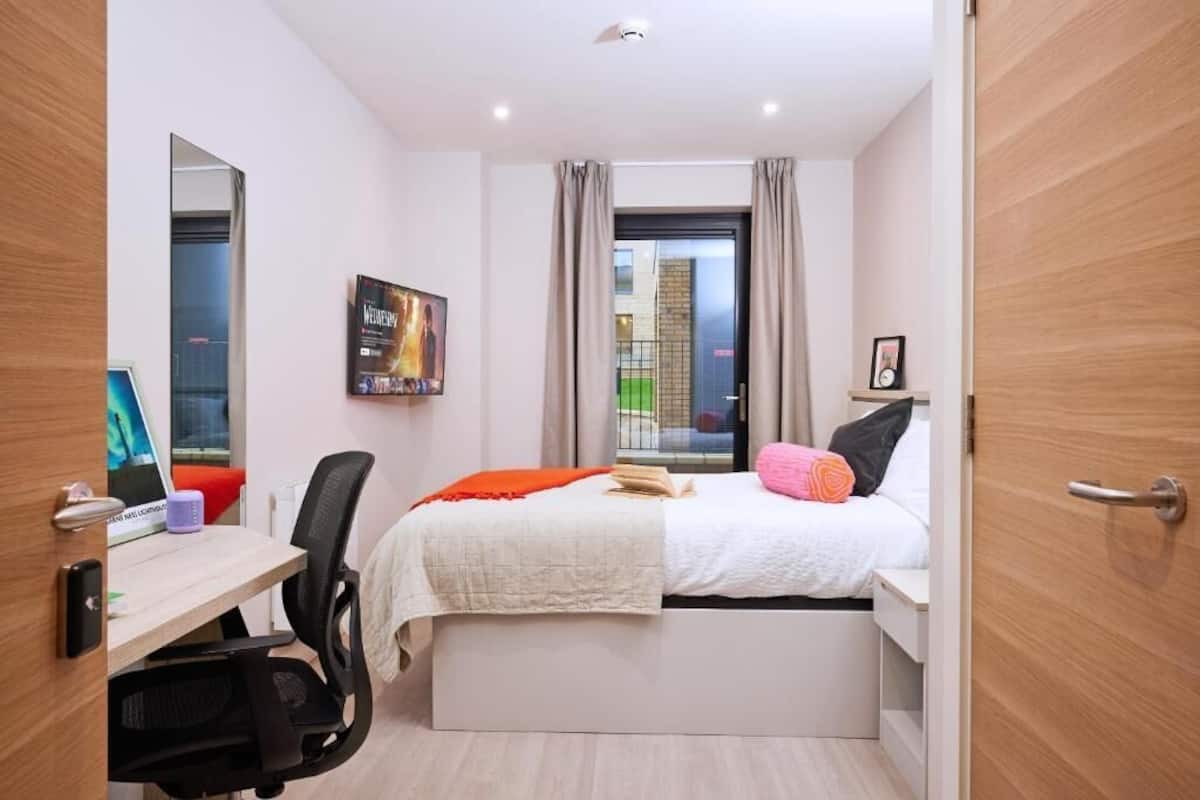A modern bedroom with a bed, desk, and chair. There is a window with curtains, a painting on the wall, and a door.