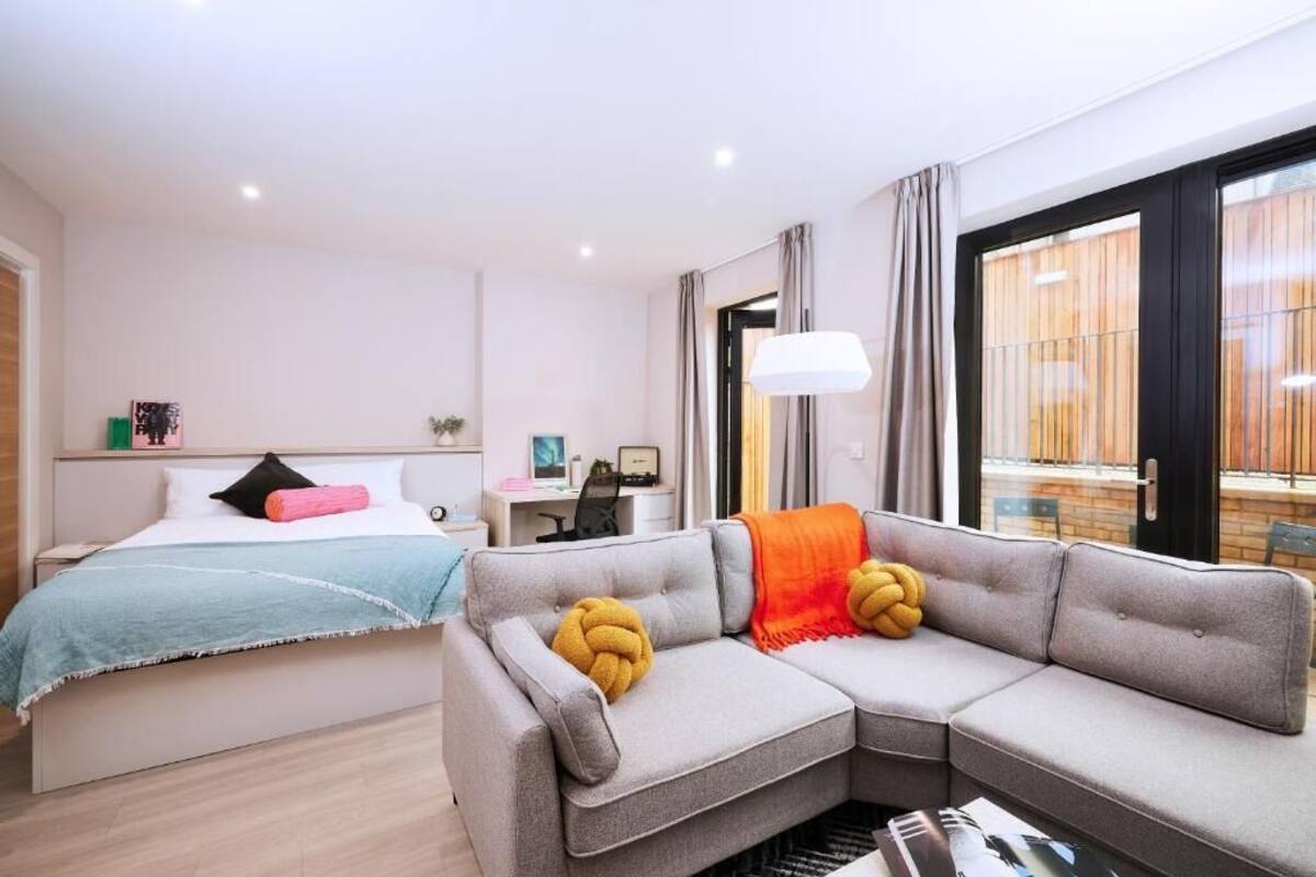 A modern hotel room with a bed, a grey sofa, a desk, and a window with curtains.
