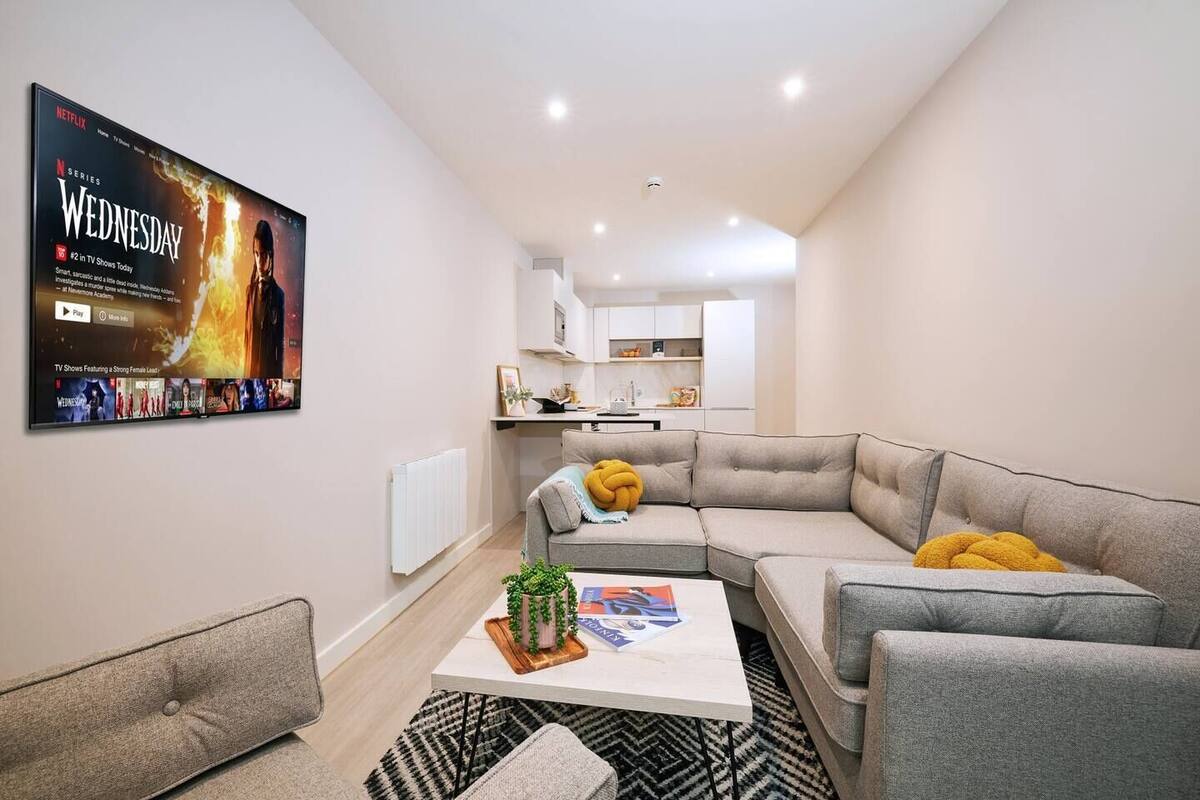 A modern living room with a grey sectional sofa, a coffee table with magazines, and a TV displaying a show. A kitchen is visible in the background.