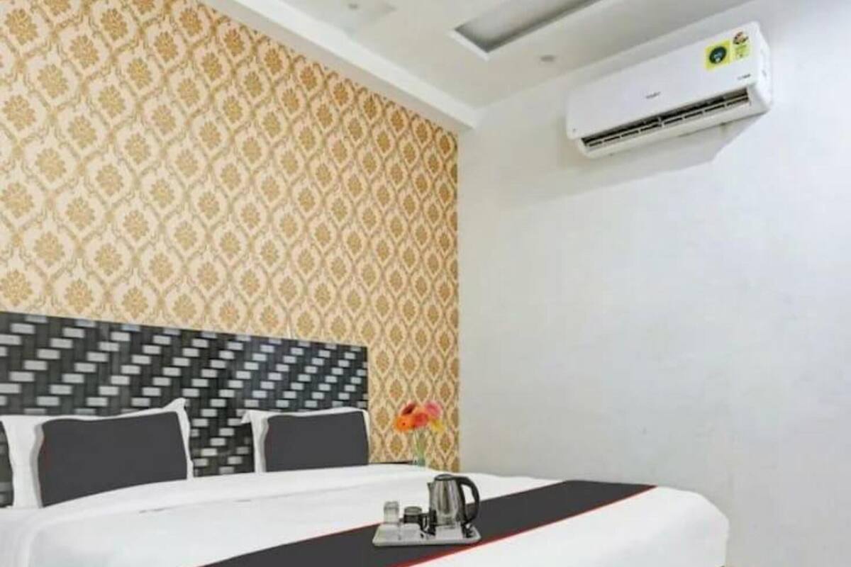 A hotel room with a patterned wallpaper, a modern bed with a black and white headboard, a small table with a kettle, and an air conditioning unit on the wall.