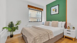 A modern bedroom with a large bed, a bedside table, a nightstand, a window with blinds, a green accent wall, and a potted plant. - Queens Quay Mansion House Flat (London)