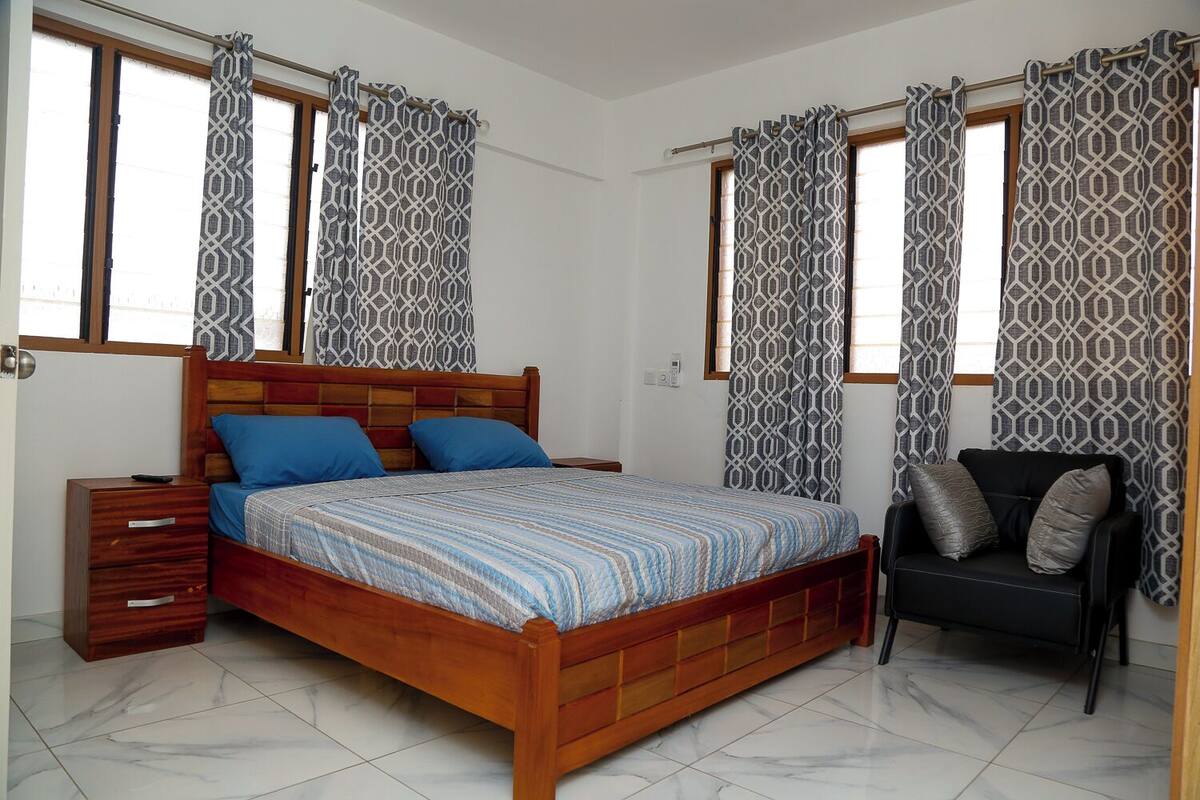 Un dormitorio con una cama de madera, dos almohadas azules, un sofá negro con cojines grises, cortinas estampadas y piso de baldosas.