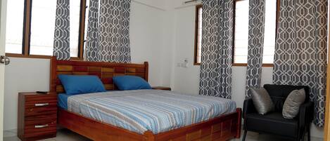 Une chambre avec un lit en bois, deux oreillers bleus, un canapé noir avec des coussins gris, des rideaux à motifs et un sol carrelé.