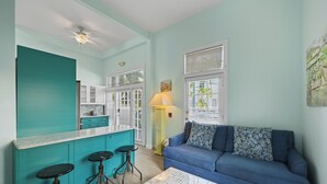 A compact living space with teal walls, a blue sofa, a kitchen area with white cabinets, and a ceiling fan.