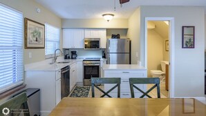A compact kitchen with white cabinets, a stainless steel refrigerator, a microwave, and a stove. There is a dining table with chairs, a ceiling fan, and a window with blinds. - Old Town Key West suite w/ pool & pet-friendly (Key West)