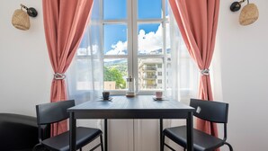 A room with a table and two chairs, a window with pink curtains, and a view of buildings and mountains. - Grande Anita - City center - Private parking (Barcelonnette)
