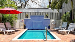 A rectangular pool with a tiled blue wall, surrounded by lounge chairs and a red umbrella.