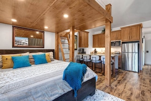 Ein Schlafzimmer mit einem Bett, Kissen, einer blauen Tagesdecke, einer Holzbalkendecke und Blick zur Küche.