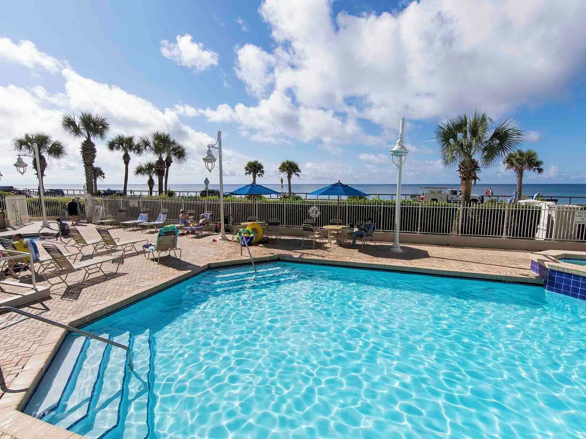 A swimming pool with clear blue water, surrounded by lounge chairs and umbrellas, with palm trees and a view of the ocean in the background.