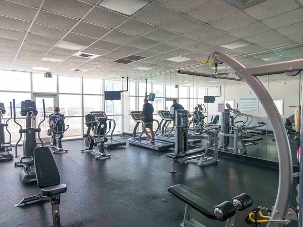 Ein Fitnessstudio mit verschiedenen Trainingsgeräten, großen Fenstern und einer Spiegelwand.