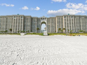 Un hôtel situé en bord de mer, doté de nombreux balcons, d’une arche centrale et d’une plage de sable devant l’établissement.