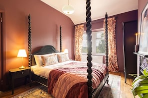 Un letto a baldacchino con una testata scura, un comodino con una lampada e una finestra con tende floreali.