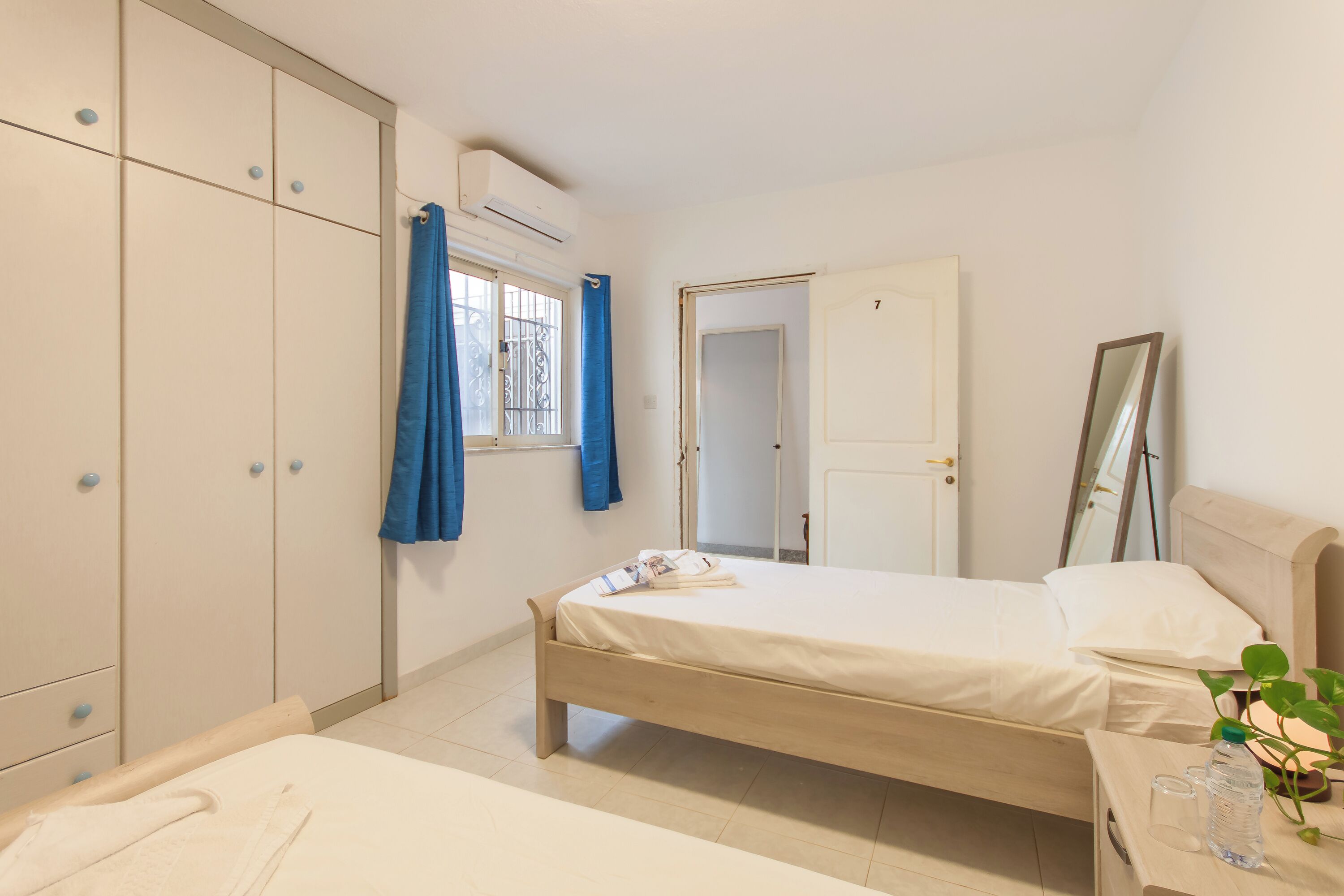 A bedroom with a bed, a wardrobe, a window with curtains, a door, and a bedside table with a bottle and glasses.