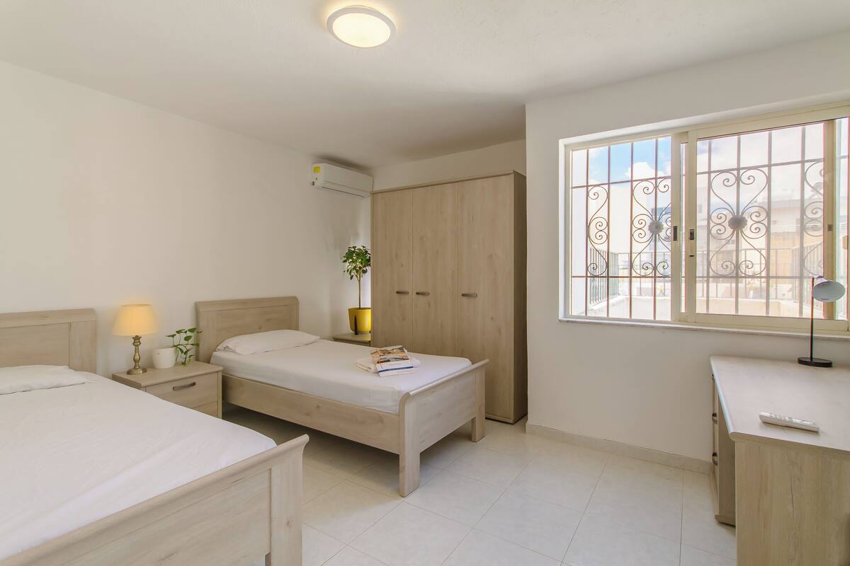 A bedroom with two beds, a desk, a wardrobe, and a window with bars.