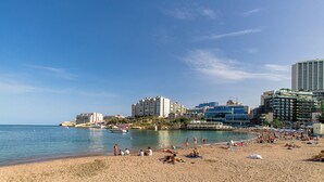 Una playa con gente tomando el sol y nadando, rodeada de edificios y un cielo despejado.