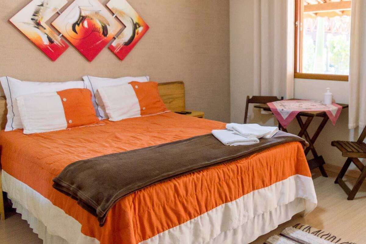 A neatly made bed with an orange and white bedspread, a brown throw blanket, and white pillows. There are two wall-mounted artworks above the bed. A small table with a pink tablecloth and a chair are visible to the right.