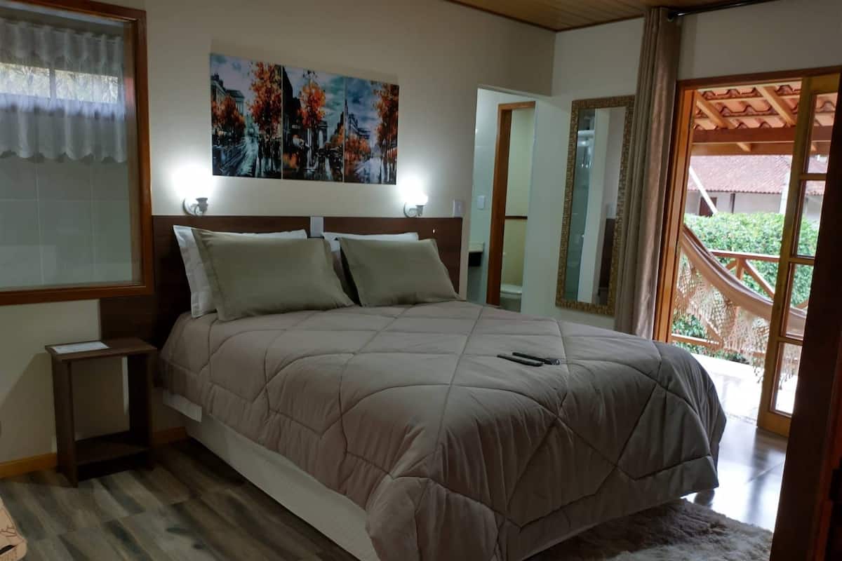 A bedroom with a large bed, a nightstand, a painting above the bed, and a view of the outdoors through a window.