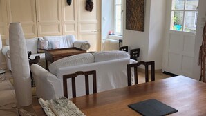 A room with a wooden dining table, white upholstered chairs, a white sofa, a white radiator, and a window with a view of greenery.