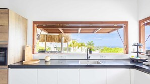 A modern kitchen with a large window, a wooden ceiling, and a view of palm trees and the ocean.