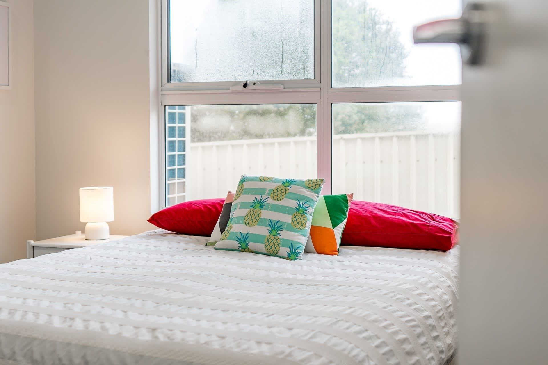 Un lit recouvert d’une courtepointe blanche à texture, un oreiller rouge, un oreiller vert orné de motifs d’ananas et une lampe blanche posée sur une table de chevet.