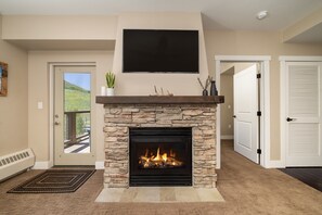 A living room with a stone fireplace, a mounted TV, and a door leading outside. - Base Camp 313 1 Bedroom Condo by RedAwning (Granby)
