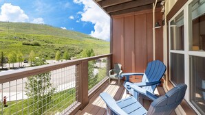 A balcony with a view of a green hillside, two Adirondack chairs, and a small table. - Base Camp 313 1 Bedroom Condo by RedAwning (Granby)