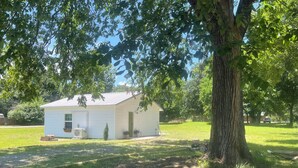 A small, single-story house with a sloped roof, a front porch, and a large tree in front.