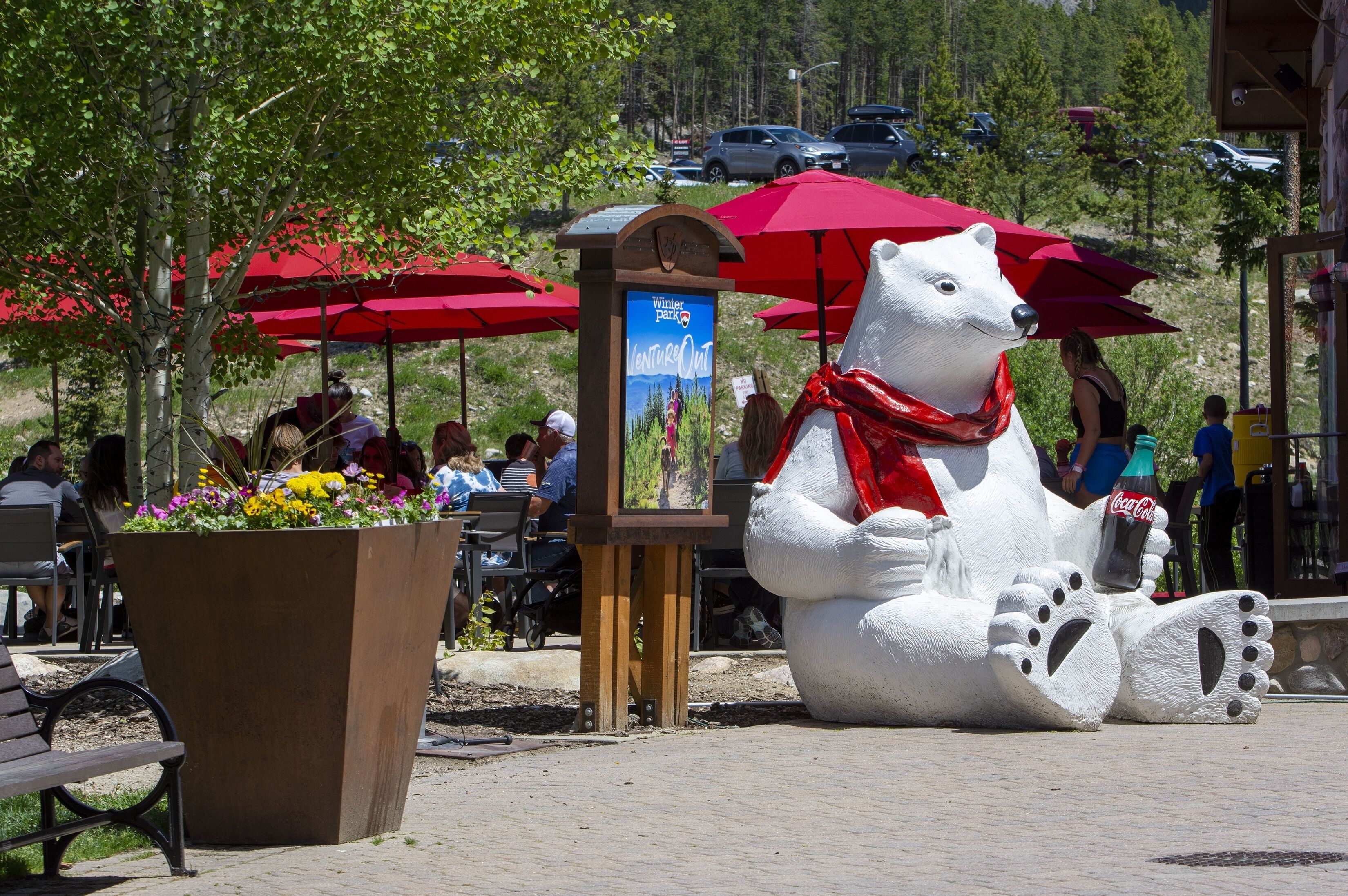 Ein öffentlicher Außenbereich mit einer großen, weißen Bärenskulptur, die eine Coca-Cola Flasche hält, umgeben von roten Sonnenschirmen und einem Schild für Winter Park.