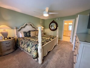 Ein Schlafzimmer mit Himmelbett, Nachttisch mit Lampe, Kommode, Deckenventilator und Spiegel an der Wand.