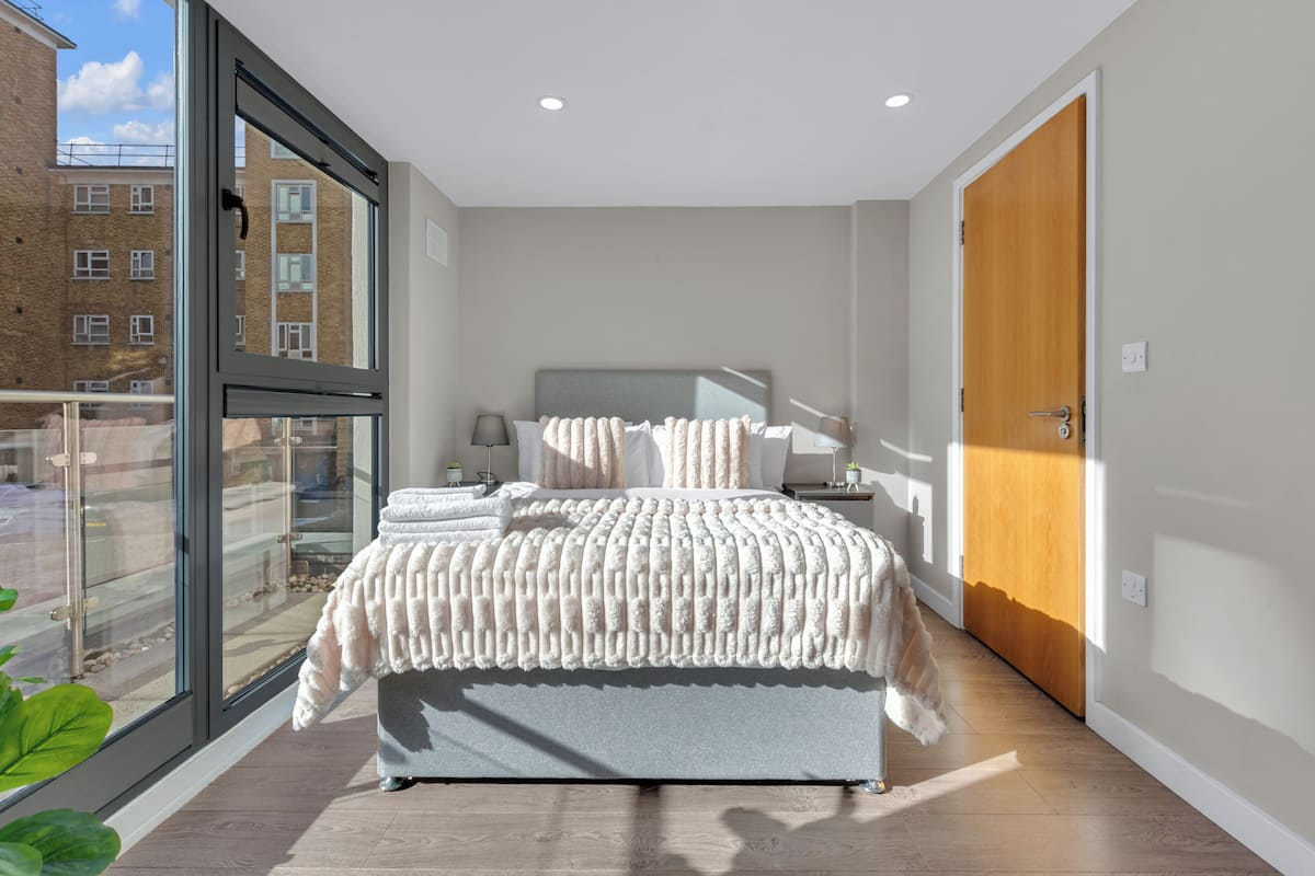 A modern bedroom with a large window, a bed with a textured blanket, a bedside table with a lamp, and a wooden door.