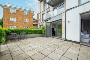 Eine moderne Wohnung mit einer Terrassenfläche, ausgestattet mit Tisch und Stühlen, mit einem Backsteinhaus im Hintergrund und einer Glastür, die zum Schlafzimmer führt.