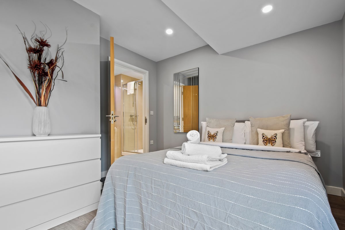 A modern bedroom with a large bed, a bedside mirror, and a vase with dried flowers on a dresser.