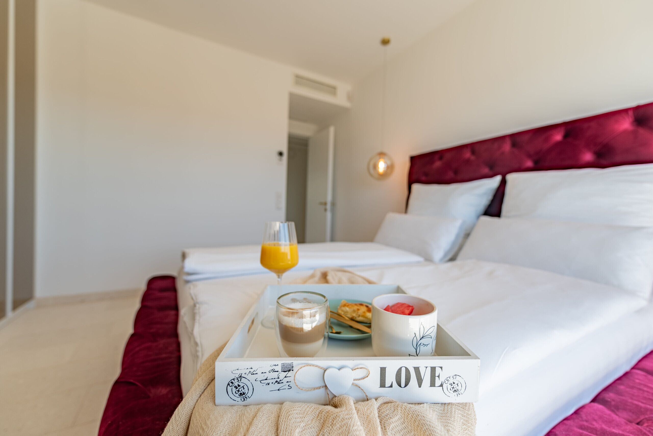Un lit bien fait, avec un plateau contenant divers éléments pour le petit-déjeuner : un verre de jus d’orange, une tasse de café, une assiette de croissants et un bol de fruits. Le lit a une tête de lit rouge et des draps blancs.