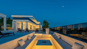 Uma área de estar moderna ao ar livre, com mesa de bilhar, assentos e guarda-corpo de vidro com vista para uma piscina.