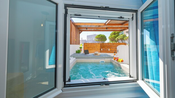 Blick durch eine Glastür auf einen Poolbereich mit Holzterrasse, Liegestuhl und Topfpflanzen.