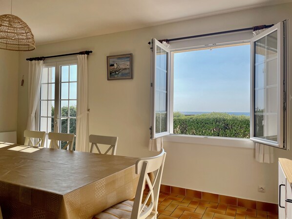 Ein Essbereich mit Tisch und Stühlen, ein Fenster mit geöffnetem Klappladen und Blick auf grüne Vegetation.