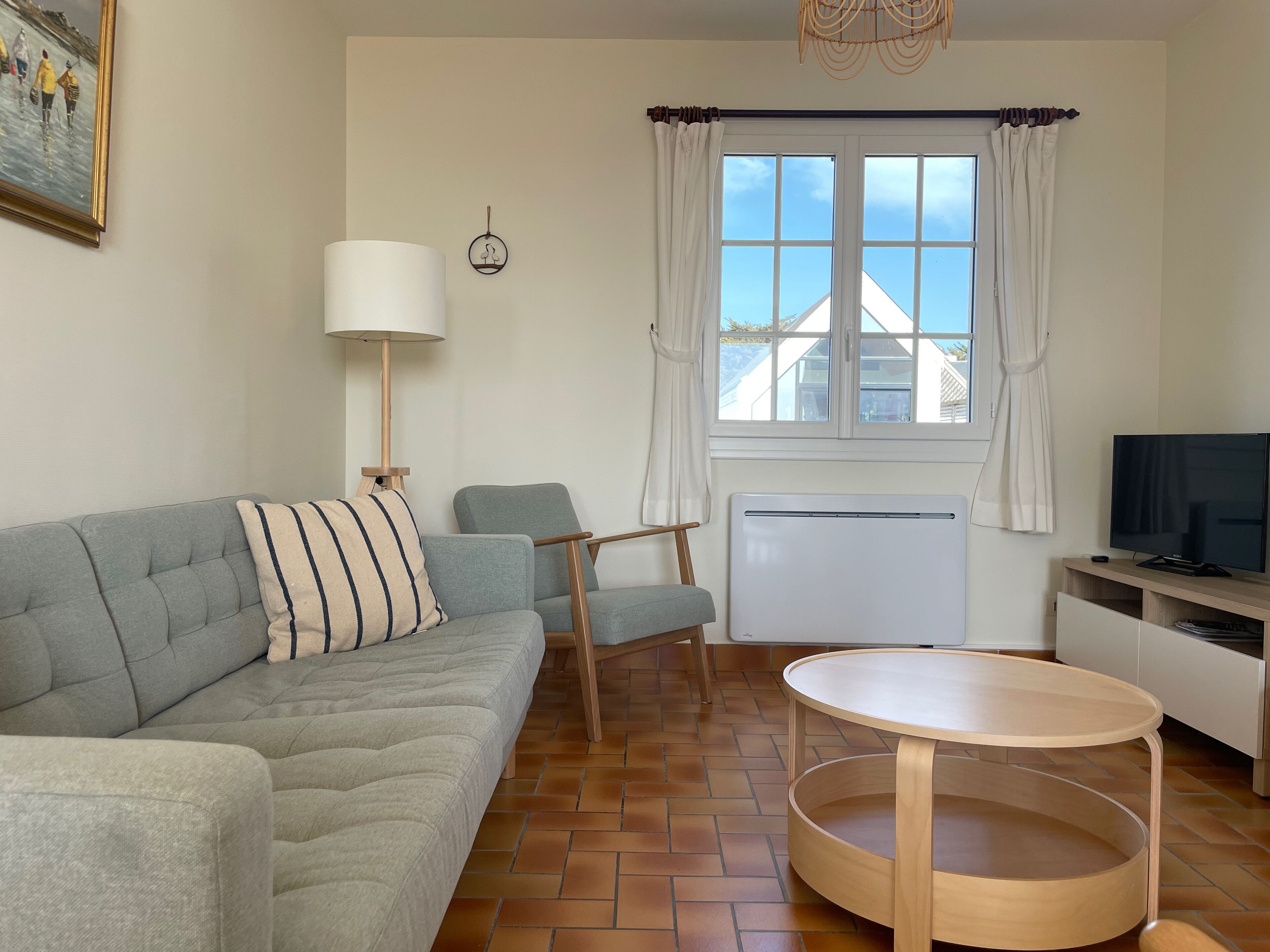Ein Wohnzimmer mit grauem Sofa, einem runden Holztisch, einem Fernseher, einer Lampe und einem Fenster mit Vorhängen.