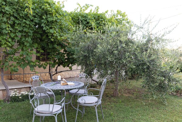 Une terrasse extérieure avec une table ronde et quatre chaises, entourée de verdure et d’un arbre.