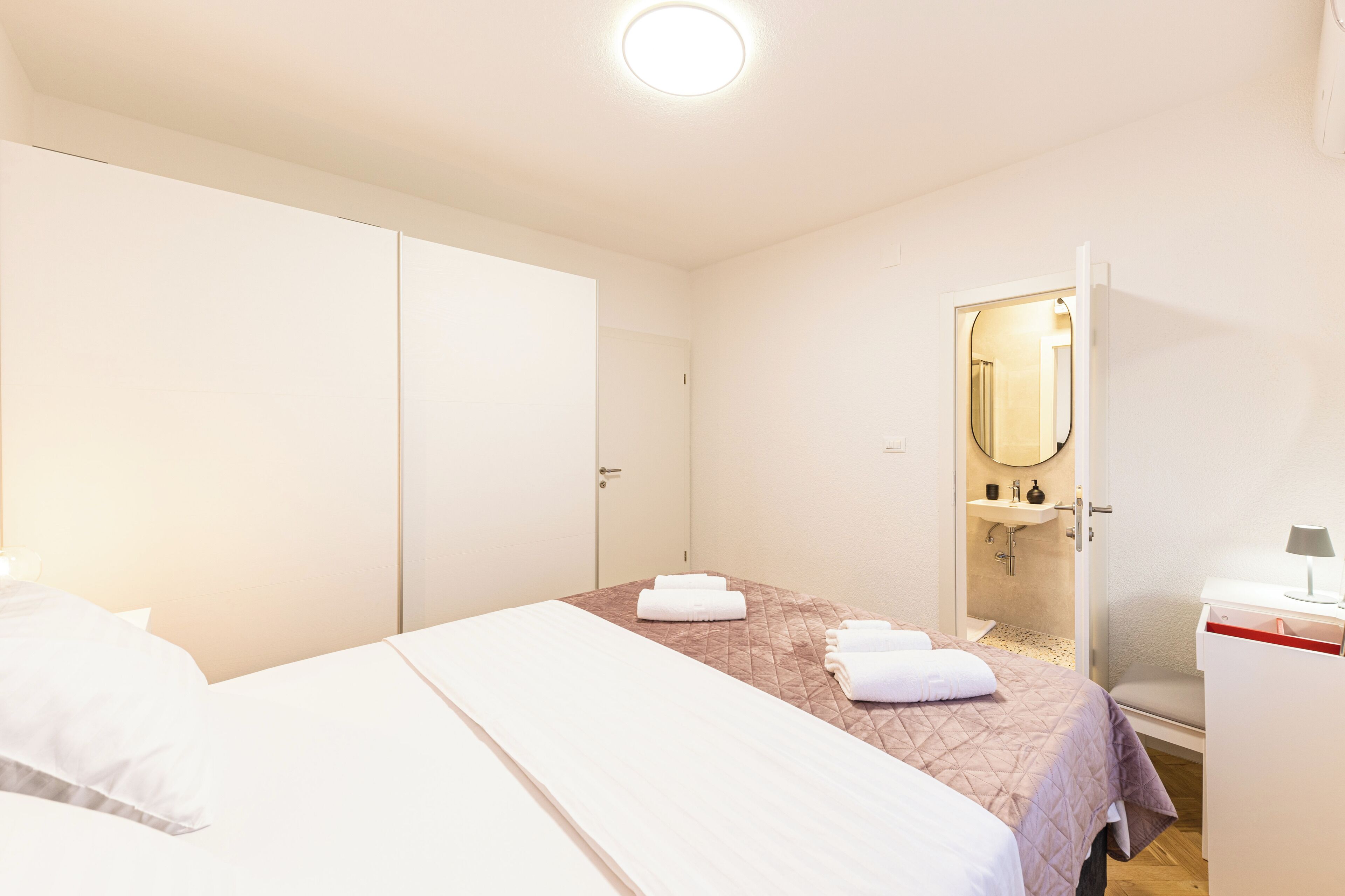 A hotel room with a bed, a bedside table, a mirror, a wardrobe, and a bathroom with a sink and mirror.