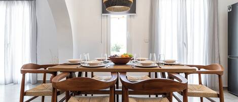 A dining area with a wooden table set for six, surrounded by woven chairs, a hanging light fixture, and a view of the outdoors through a window.