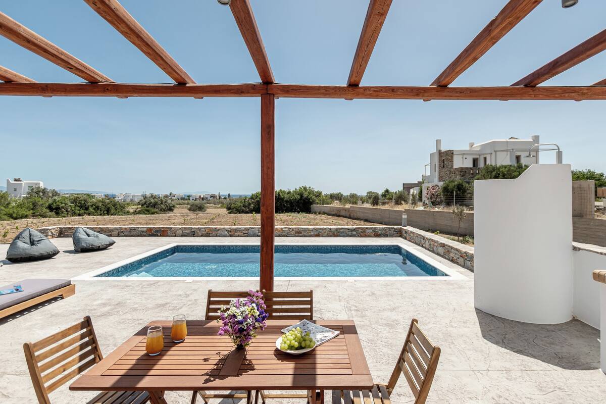 Una pèrgola de fusta sobre una zona de piscina amb una taula preparada per a dos, un vas de flors i un bol de fruita.