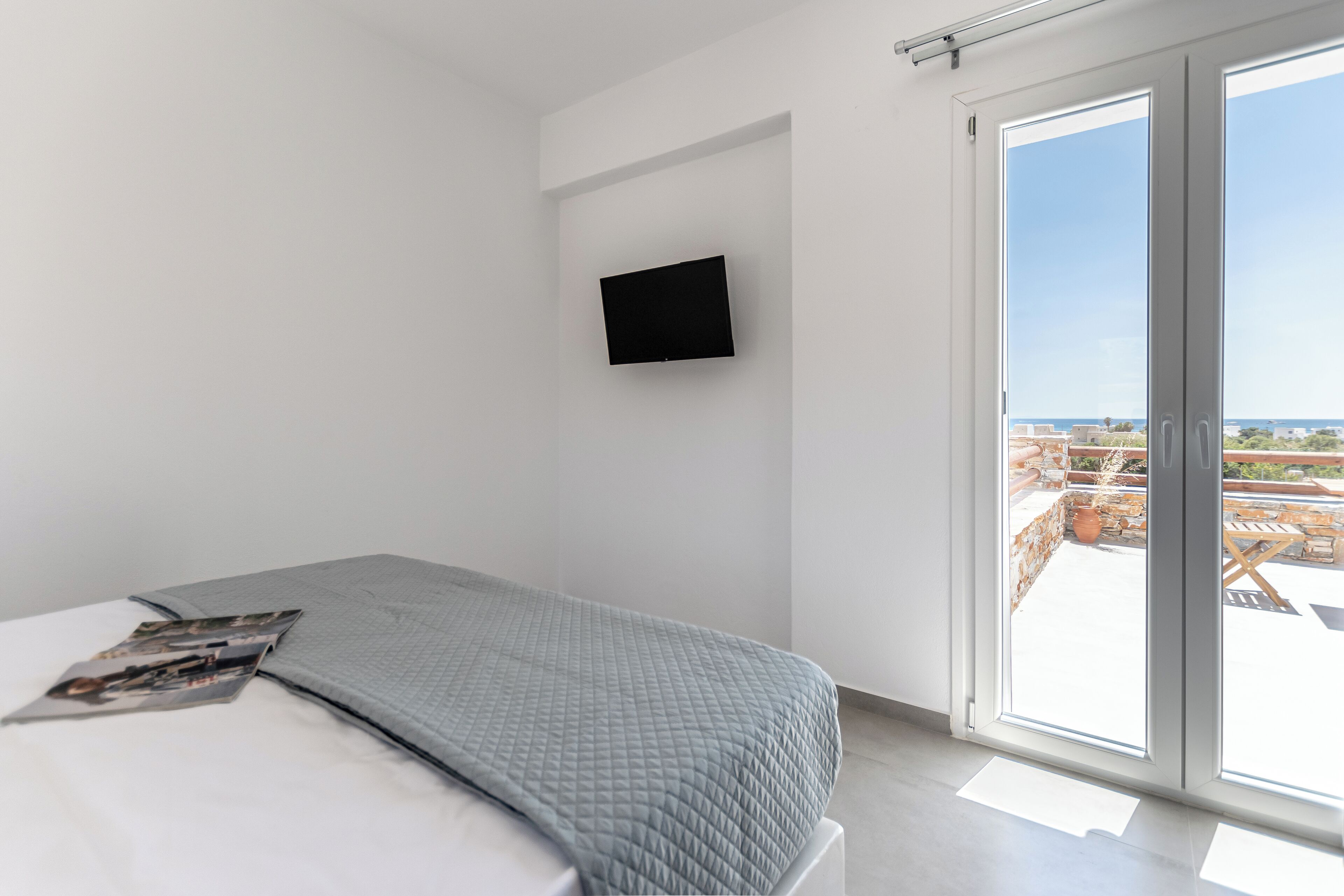 A bedroom with a bed, a flat-screen TV mounted on the wall, and a sliding glass door leading to an outdoor balcony.
