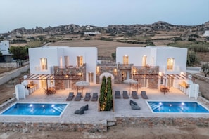 Una villa moderna con dos piscinas, mobiliario de exterior y vistas a colinas rocosas.