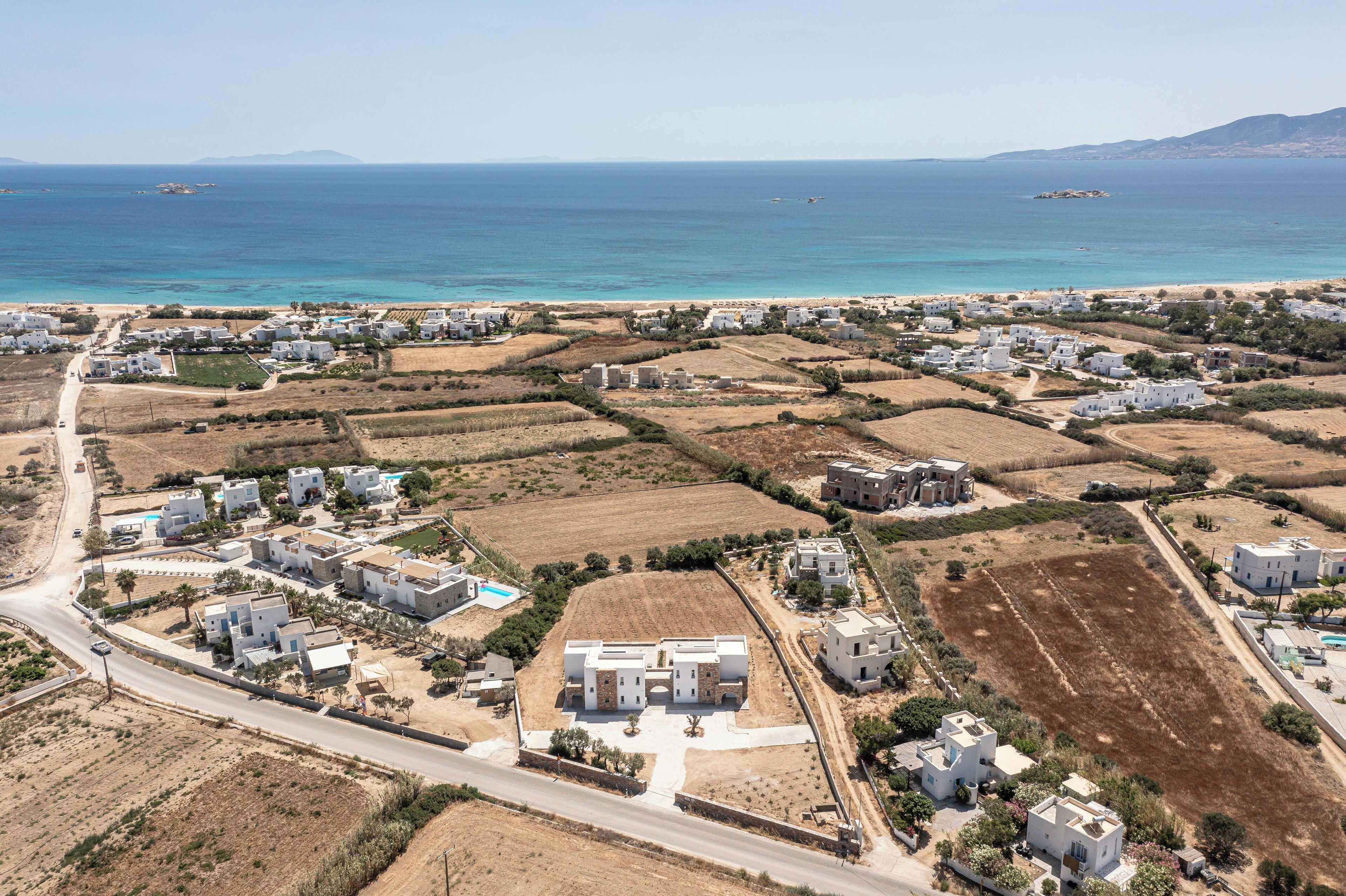 Vue aérienne d’un quartier résidentiel situé sur la côte, avec des bâtiments blancs, une route et une plage.