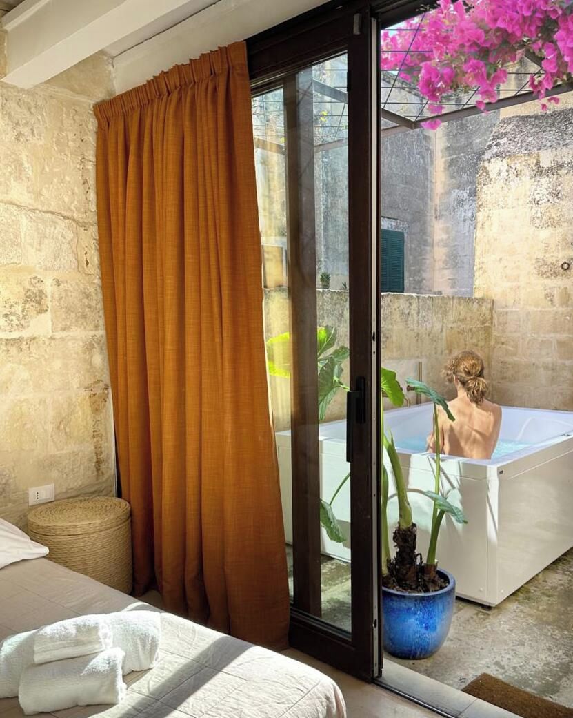 A room with a bed, a large window with curtains, a sliding glass door leading to a balcony, and a bathtub.