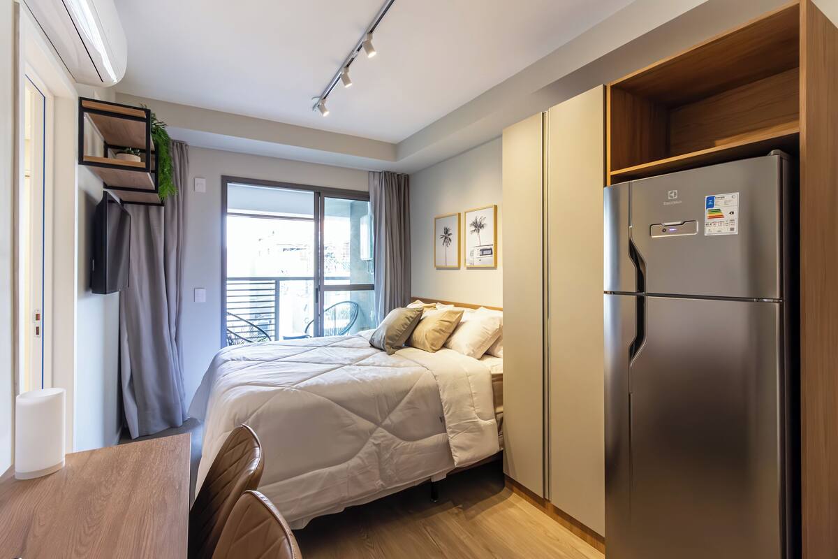 Une chambre d’hôtel de taille réduite, comprenant un lit, un réfrigérateur, un coin repas et une fenêtre avec des rideaux.