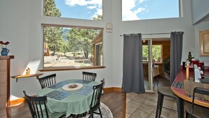 Un coin repas avec une table dressée pour quatre personnes, de grandes fenêtres offrant une vue sur l’extérieur et un espace cuisine avec un comptoir et des appareils électroménagers.