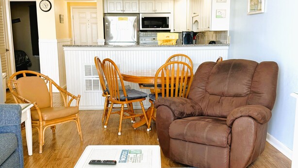 Een woonkamer met een bruine leren bank, houten stoelen, een eettafel en een keuken op de achtergrond.
