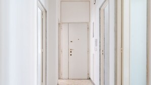 Um corredor estreito com uma porta branca no final, ladeado por paredes brancas e piso de azulejos.